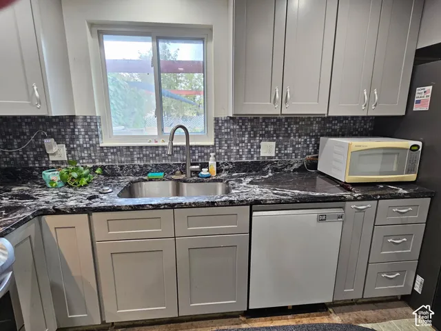 Kitchen featuring grey shaker-style cabinetry, high-contrast dark stone countertops, a mosaic tile backsplash, stainless steel double-basin sink with an arched pull-down faucet, and a built-in dishwasher