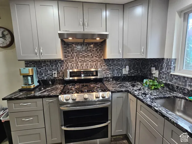 Shaker-style cabinetry, dark stone-patterned countertops, stainless steel gas range, integrated range hood, and a decorative mosaic tile backsplash