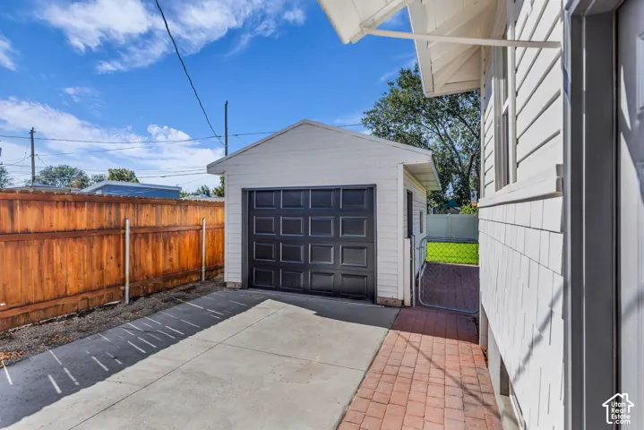 Detached garage featuring white siding, a black paneled door, concrete driveway, and red brick paver walkway