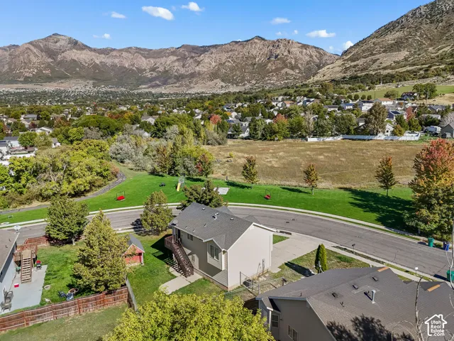 Aerial perspective featuring a two-story residence, asphalt shingle roofing, concrete driveway, and a wooden exterior deck, set against a backdrop of prominent mountain terrain