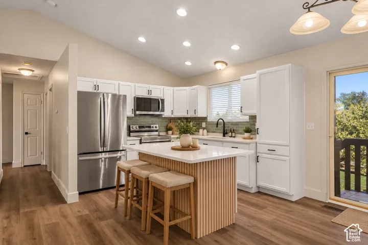Kitchen featuring white shaker cabinetry, a center island with a vertical-slat wood-finish base, stainless steel appliances, a textured tile backsplash, and wood-finish flooring