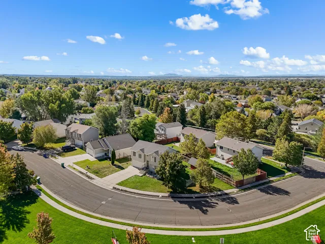 Aerial perspective of a corner lot featuring a residential structure with an attached multi-car garage, concrete driveway, fenced perimeter, and mature landscape elements
