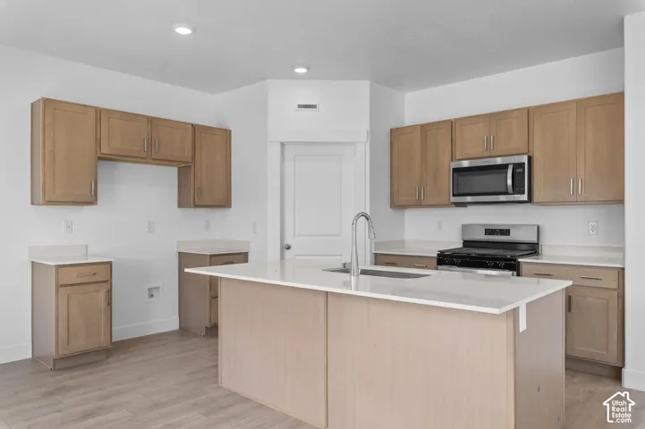 Kitchen featuring light wood-finish cabinetry, quartz countertops, a central island with an integrated sink, stainless steel microwave, and wood-finish flooring