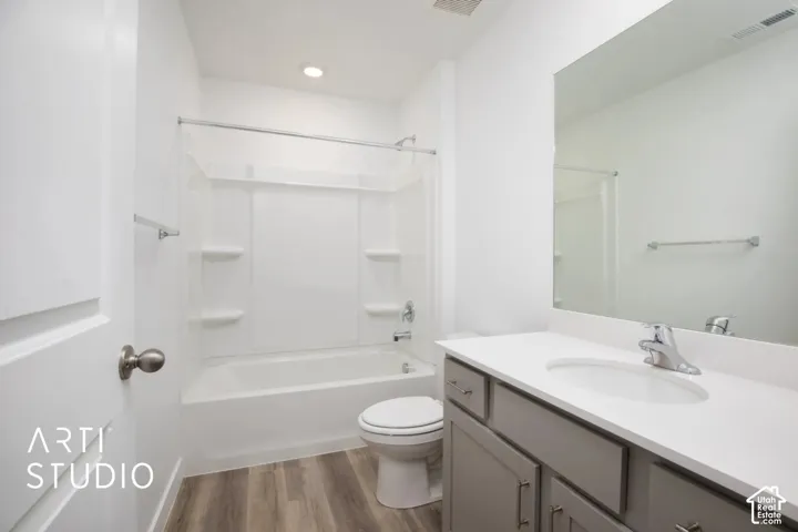 Full-width mirror, vanity with a light-toned solid surface countertop, white tub-shower combination with integrated shelving, and wood-finish flooring