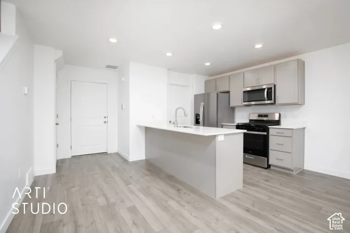 Kitchen featuring gray shaker-style cabinetry, white countertops, stainless steel appliances, a center island with a dual-basin sink, and wood-finish flooring