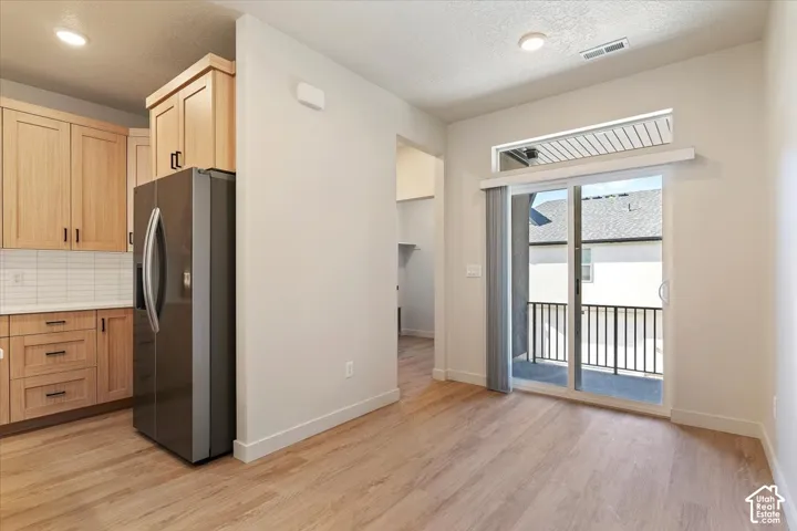 Kitchen featuring light wood-finish cabinetry, white subway tile backsplash, stainless steel refrigerator, wood-finish flooring, and a sliding glass door with a transom window