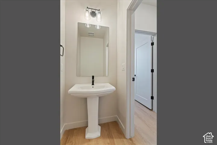 Pedestal sink with a tall black faucet, frameless wall mirror, two-light fixture with glass shades, and wood-finish flooring