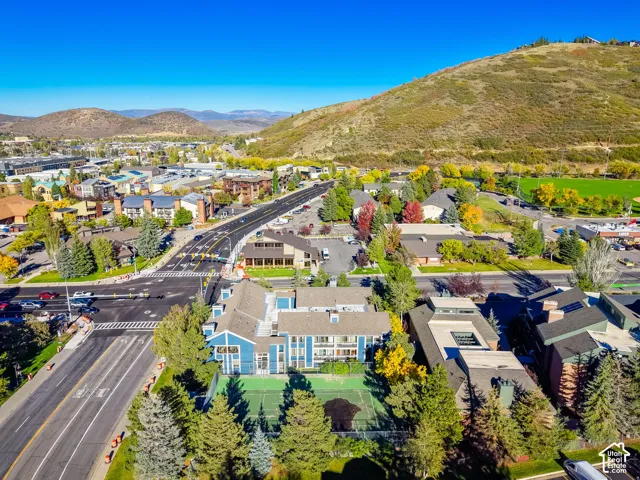 Aerial perspective of a multi-level residential building featuring blue siding, a dedicated tennis court, mature evergreen landscaping, and a mountainous backdrop
