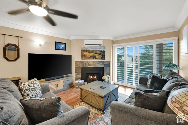 Corner fireplace with a stacked-stone surround, wood-finish flooring, plantation shutters, wall-mounted ductless climate control unit, and a ceiling fan with integrated lighting