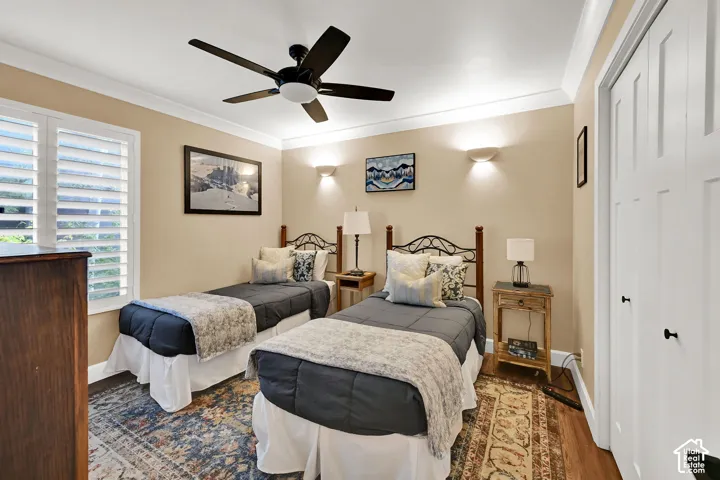 Bedroom featuring wood-finish flooring, crown molding, plantation shutters, a ceiling fan, and dual wall-mounted sconces