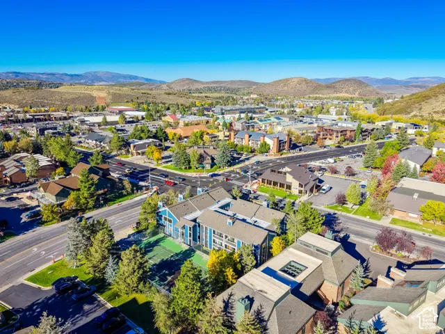 Aerial perspective of a multi-story building complex featuring a fenced-in tennis court, mature evergreen trees, surface parking, and a backdrop of mountain terrain