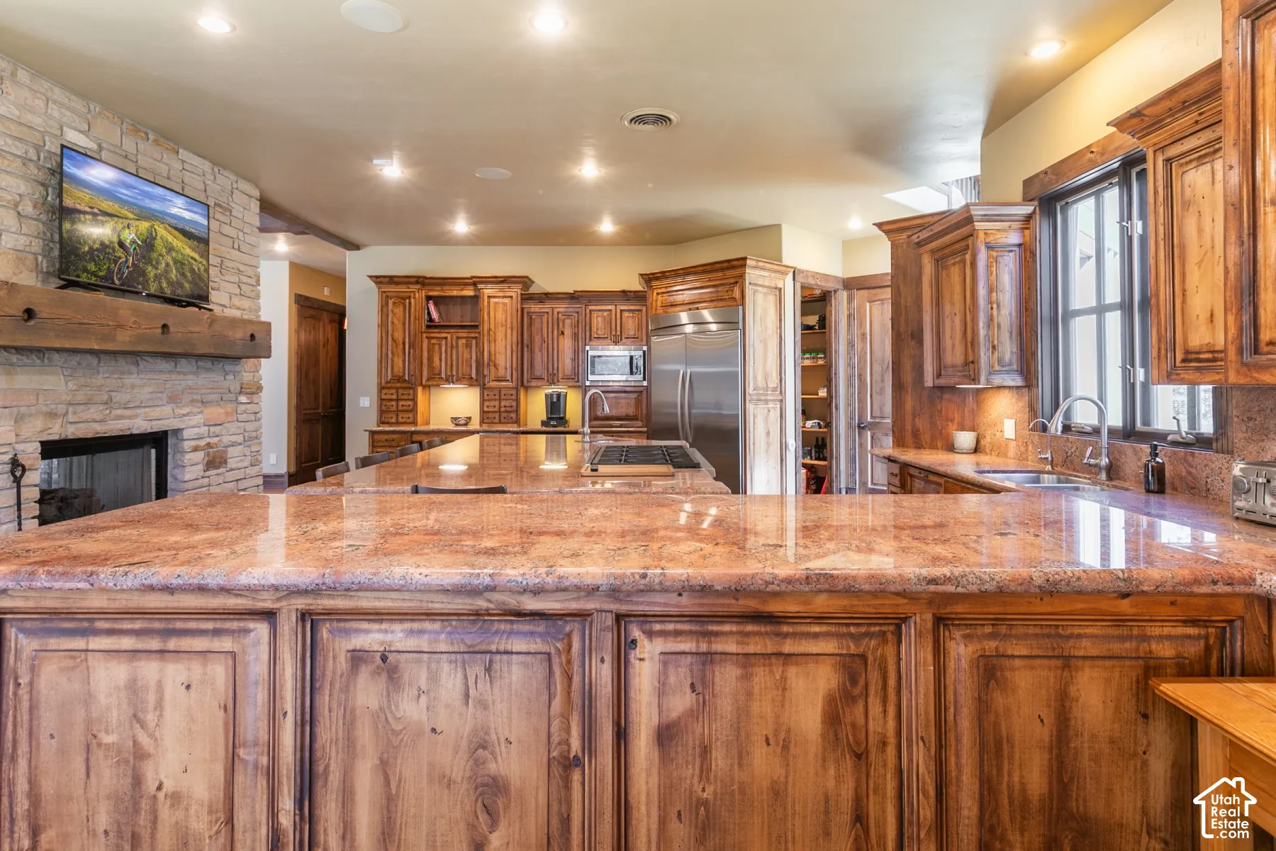 Kitchen featuring custom wood cabinetry, expansive stone countertops, stainless steel appliances, stone fireplace surround, and recessed lighting