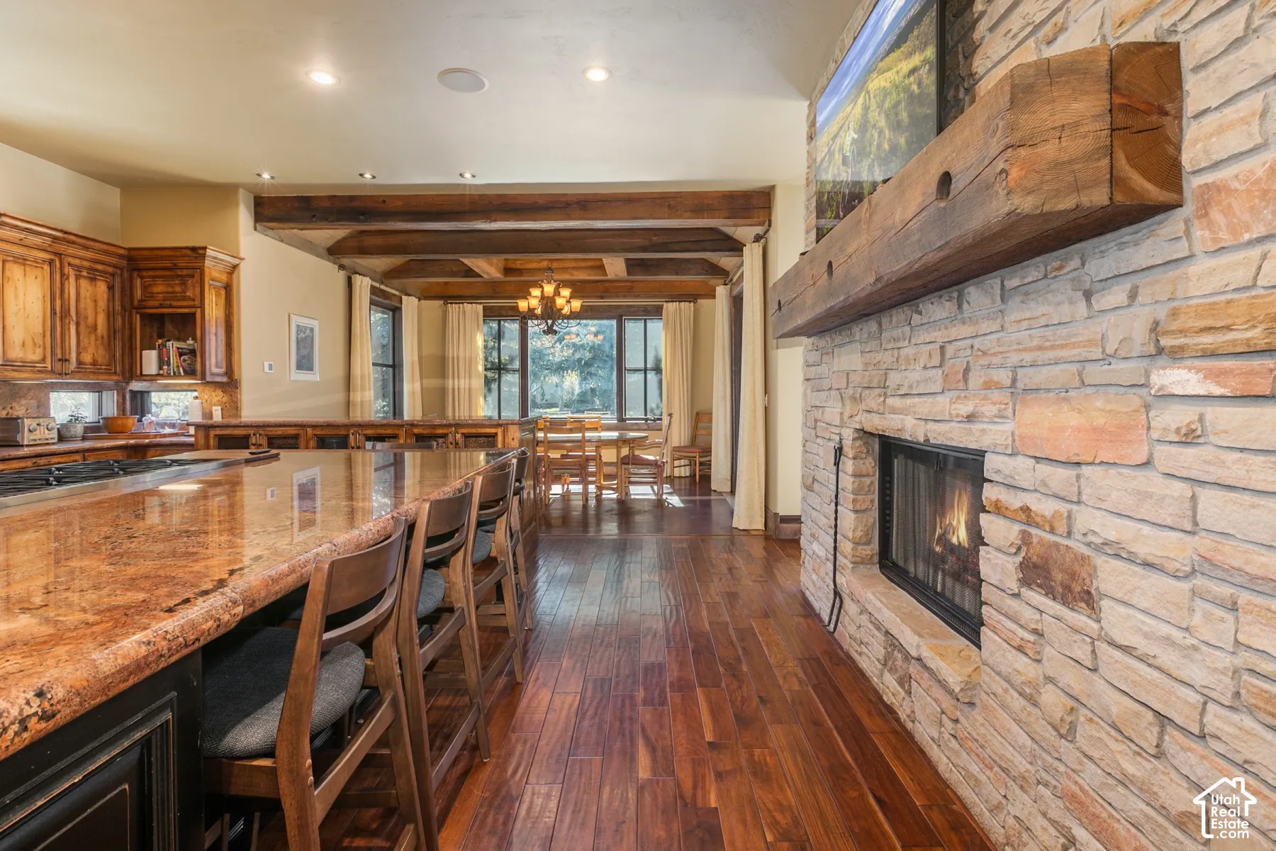 Stone fireplace with a prominent timber mantel, hardwood flooring, exposed wood ceiling beams, and stone-finished kitchen island