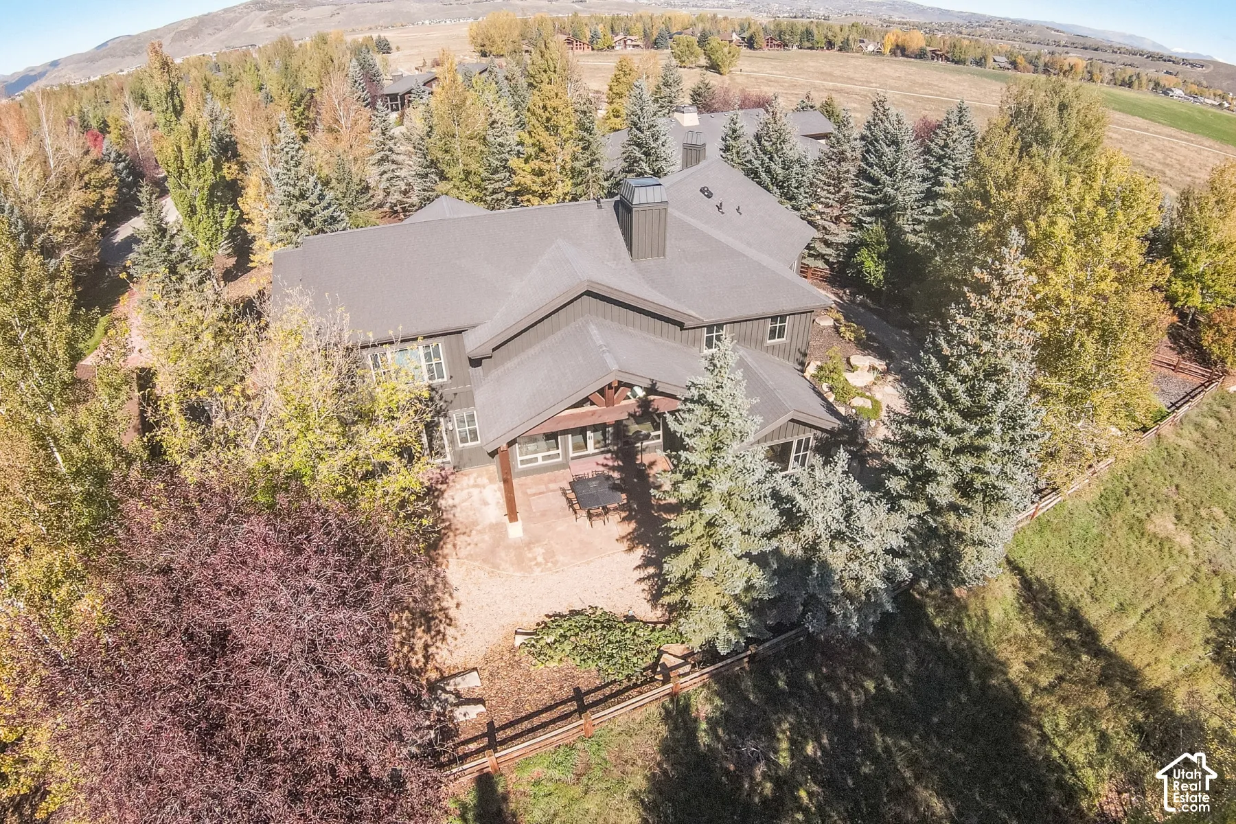 Aerial view of a multi-level residence with dark vertical siding, a metal roof, prominent chimney stack, stone-surfaced patio, and timber-frame gable accents