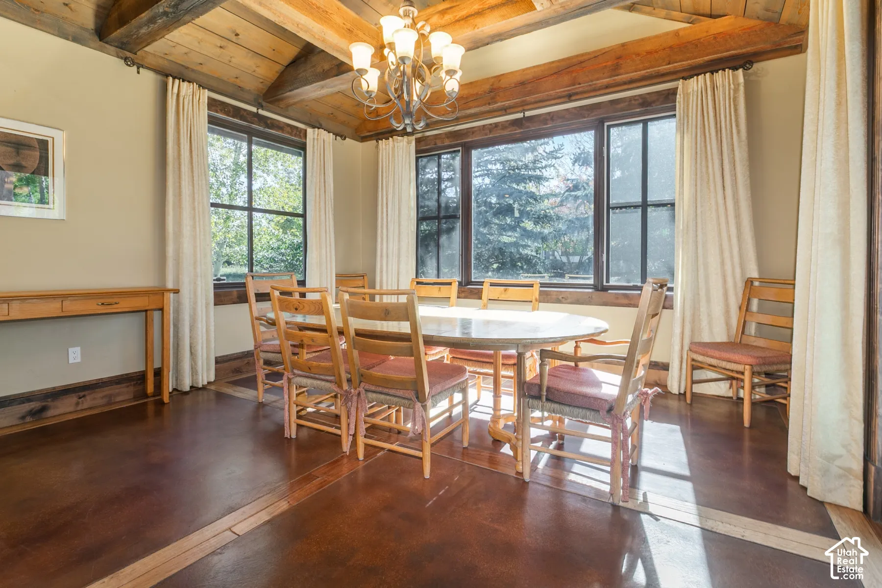 Vaulted ceiling featuring exposed timber beams, stained concrete flooring with wood inlay borders, multi-pane windows, and a classic metal chandelier