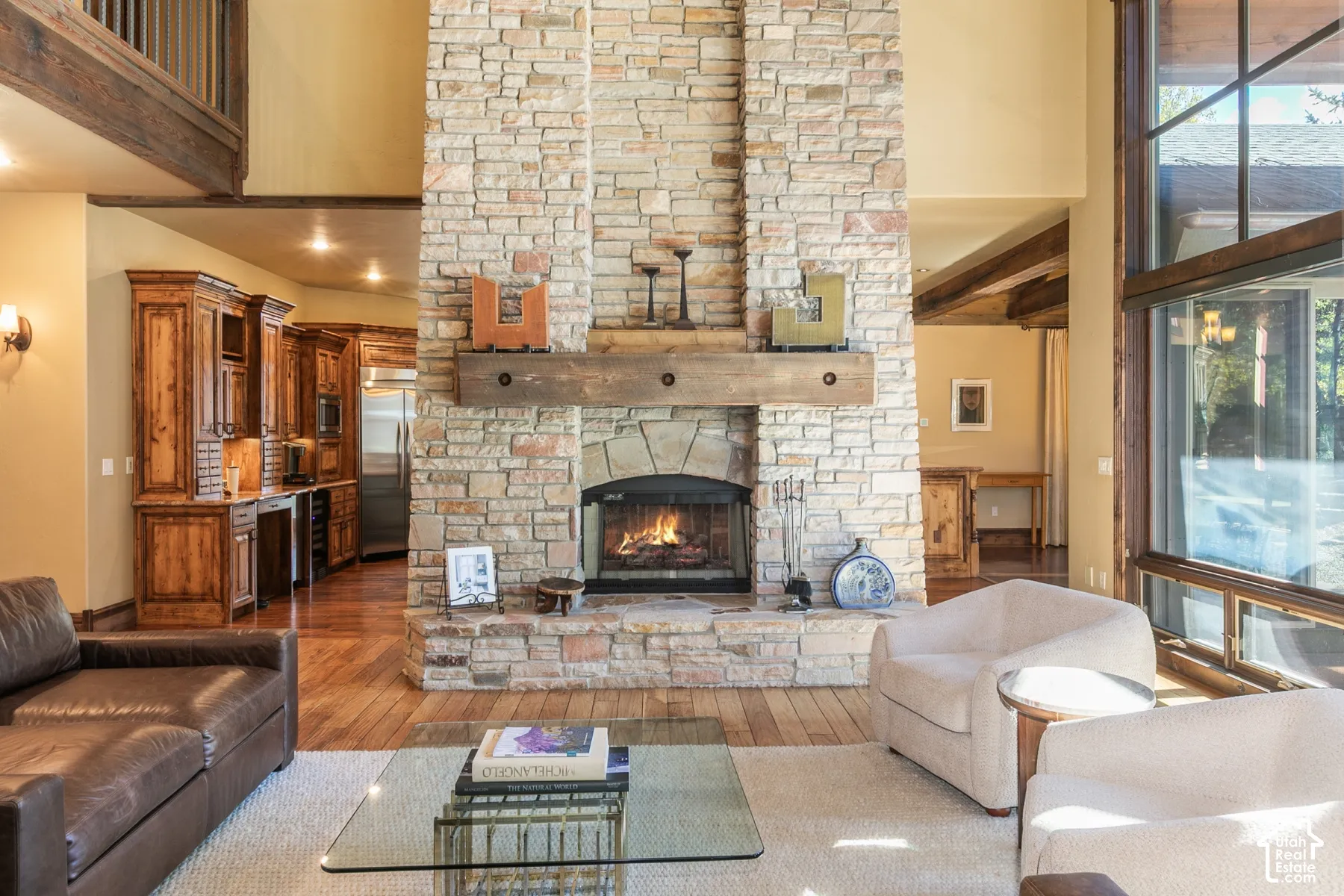 Double-height floor-to-ceiling stone fireplace with a substantial timber mantel, wood-finish flooring, extensive built-in cabinetry, and expansive multi-pane windows