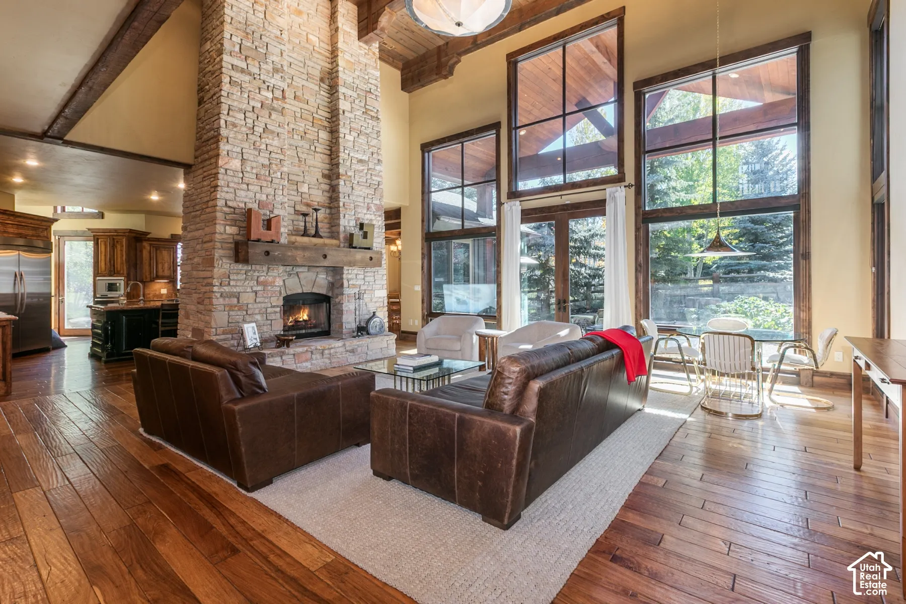 Floor-to-ceiling stone fireplace with a timber mantel, soaring vaulted ceilings with exposed wood beams, expansive multi-pane windows, and wide-plank wood-finish flooring