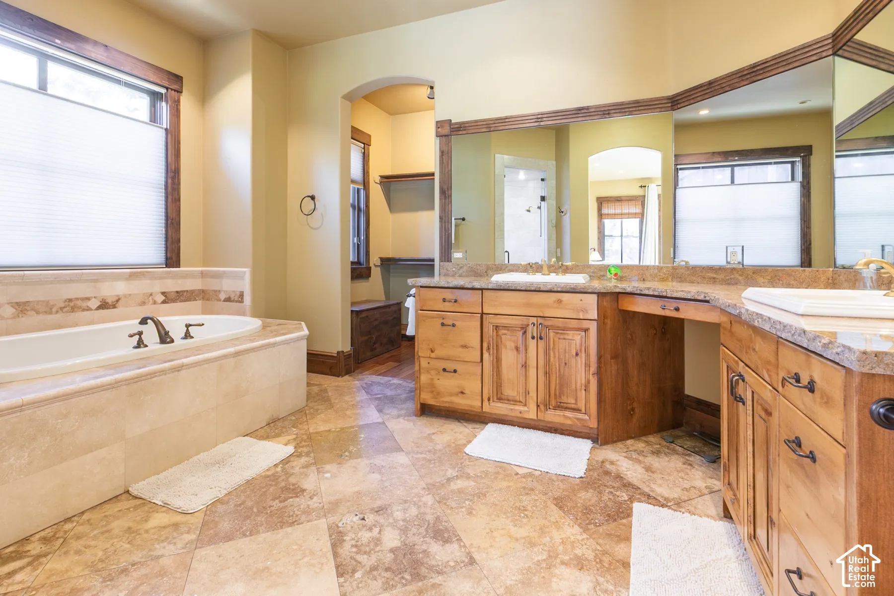 Natural stone tile flooring, custom wood cabinetry with integrated vanity, drop-in soaking tub with stone tile surround, stone countertops, and large wall-mounted mirrors