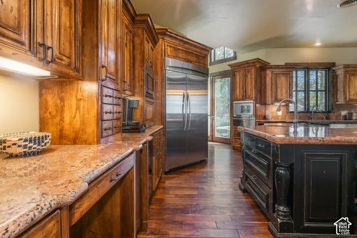 Custom wood cabinetry, stone countertops, wood-finish flooring, built-in stainless steel appliances, and a black finish kitchen island with decorative pillar detailing