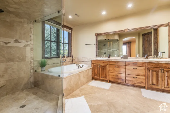 Dual-sink vanity with natural wood cabinetry, stone-surround soaking tub, glass-enclosed walk-in shower, stone tile flooring, and large framed wall mirror