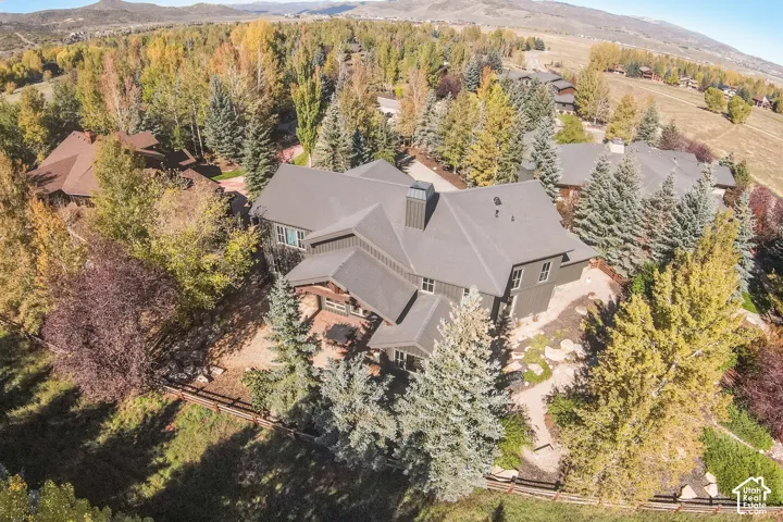 Aerial perspective of a multi-gabled residence featuring vertical siding, a composition shingle roof, a stone-accented exterior, and a stone-paved patio surrounded by mature evergreen trees