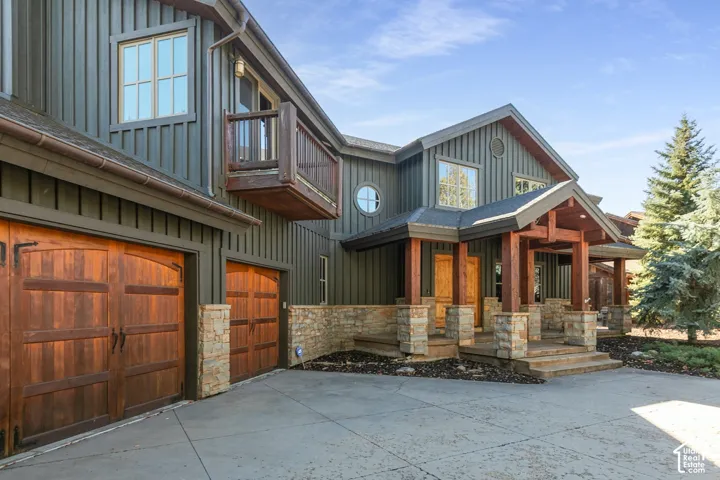 Exterior featuring vertical wood-style siding, stone veneer accents, timber-frame covered entryway, wooden carriage-style garage doors, and a second-story balcony
