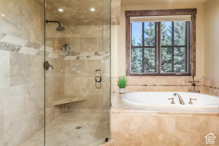 Walk-in shower with stone tile surround, built-in corner bench, and frameless glass door, accompanied by an oversized jetted tub featuring a stone tile deck and wood-trim window casing