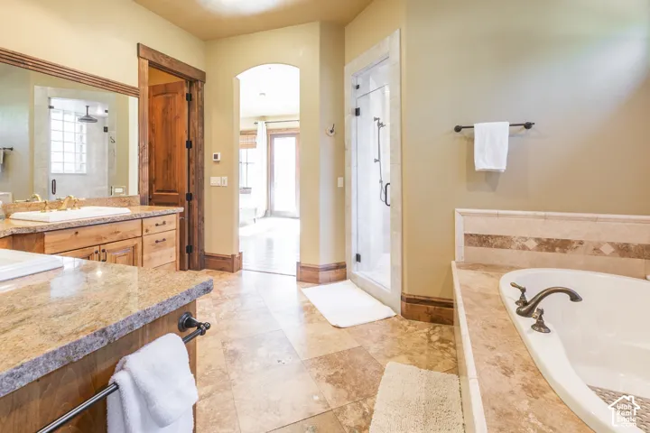 Bathroom featuring a stone-topped dual vanity with wooden cabinetry, a jetted soaking tub with tile surround, a glass-enclosed shower, and tile flooring