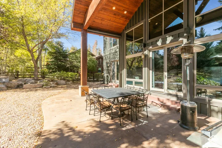Covered stone patio featuring a timber-framed overhang, expansive floor-to-ceiling windows, and natural landscaping with rock outcroppings