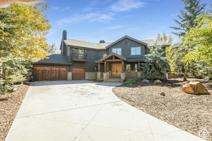 Two-story exterior featuring vertical siding, stone veneer accents, wood-paneled garage doors, a covered timber-framed entry, and a second-story balcony