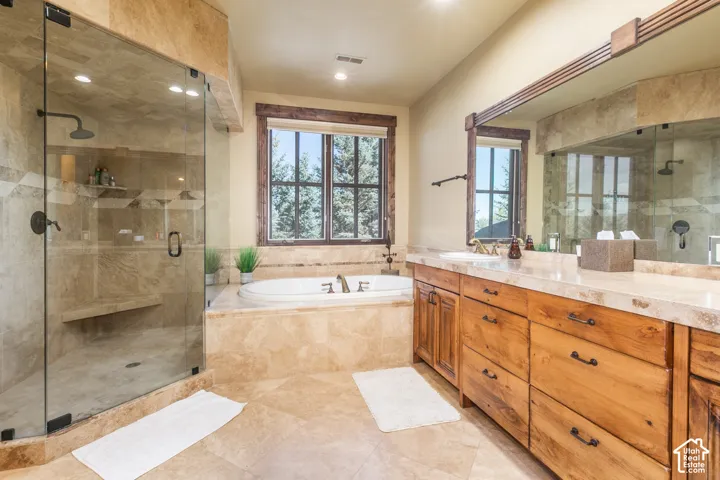 Bathroom featuring stone tile flooring and surrounds, a glass-enclosed shower with built-in seating, a deep soaking tub, wood cabinetry with a stone countertop, and a large wall-mounted mirror
