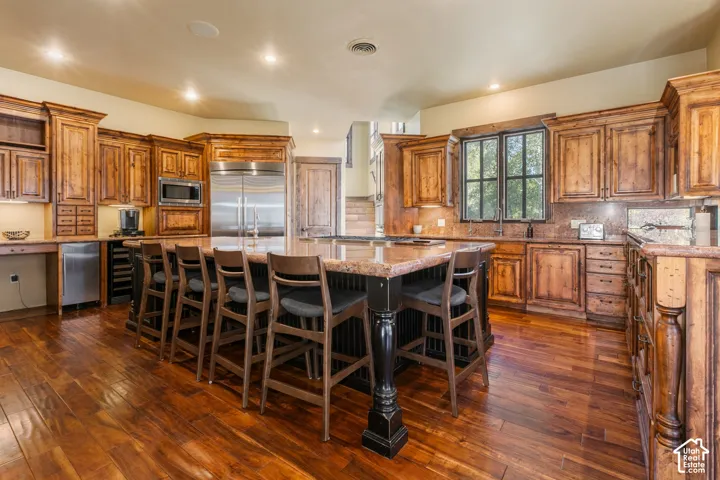 Kitchen featuring custom knotty wood cabinetry, stone countertops, a large central island with seating, built-in stainless steel appliances, and wood flooring