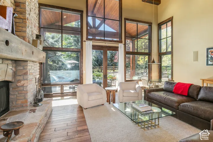 Double-height living area featuring floor-to-ceiling windows, exposed timber ceiling beams, full-height stone masonry fireplace with a timber mantel, and wood-finish flooring