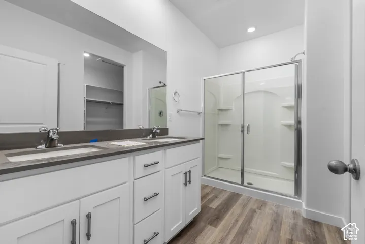 Dual-sink vanity with a solid-surface countertop, white cabinetry featuring matte hardware, wood-finish flooring, and a walk-in shower with a glass enclosure