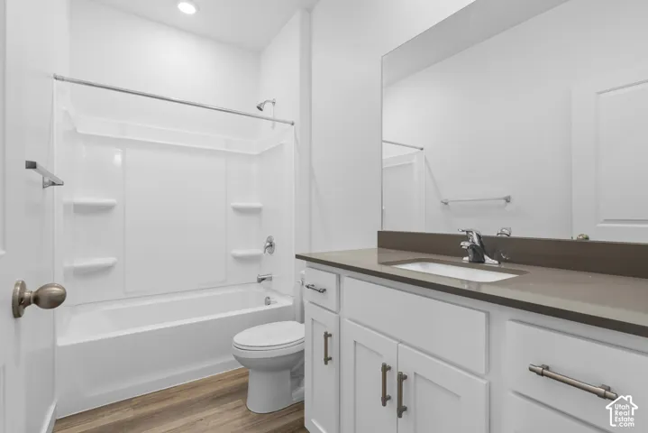 Bathroom featuring white shaker-style cabinetry, dark solid-surface countertop, rectangular undermount sink, and wood-finish flooring, accompanied by a bathtub-shower combination with integrated shelving