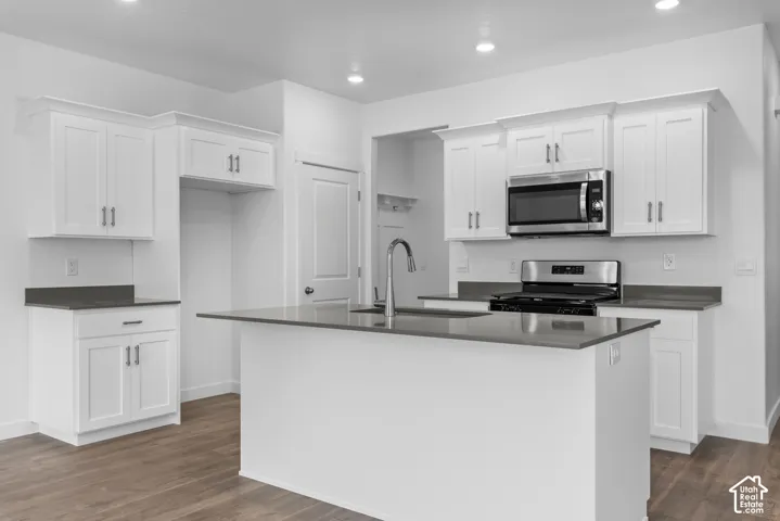 Kitchen featuring white shaker-style cabinetry, solid-surface countertops, a center island with an undermount sink, stainless steel appliances, and wood-finish flooring