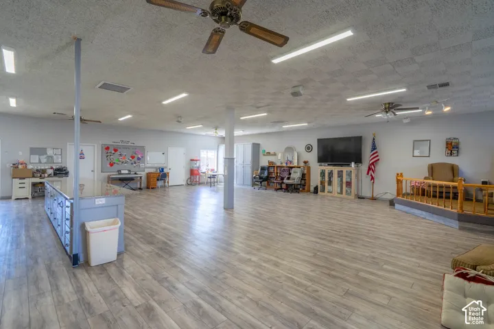 Expansive open-concept room featuring wood-finish flooring, acoustic ceiling tiles, integrated overhead lighting, ceiling fans, and a raised platform with wooden balustrade