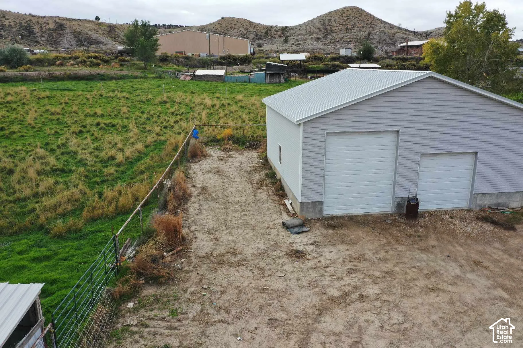 Detached garage featuring light-colored horizontal siding, dual white metal roll-up doors, and a metal roof, set on a multi-acre parcel with perimeter wire fencing and mountain topography