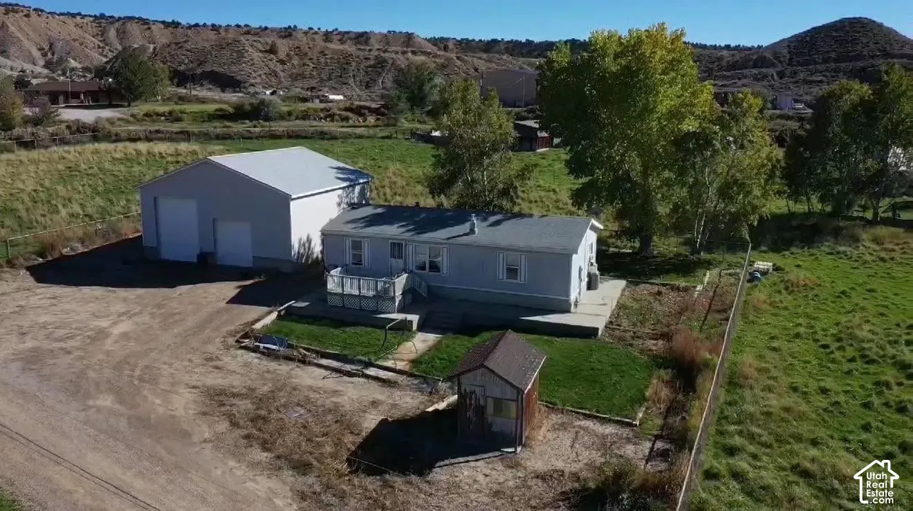 Detached metal-sided garage, raised wooden deck, concrete patio, and small accessory structure set on a parcel with mature trees and mountain surroundings