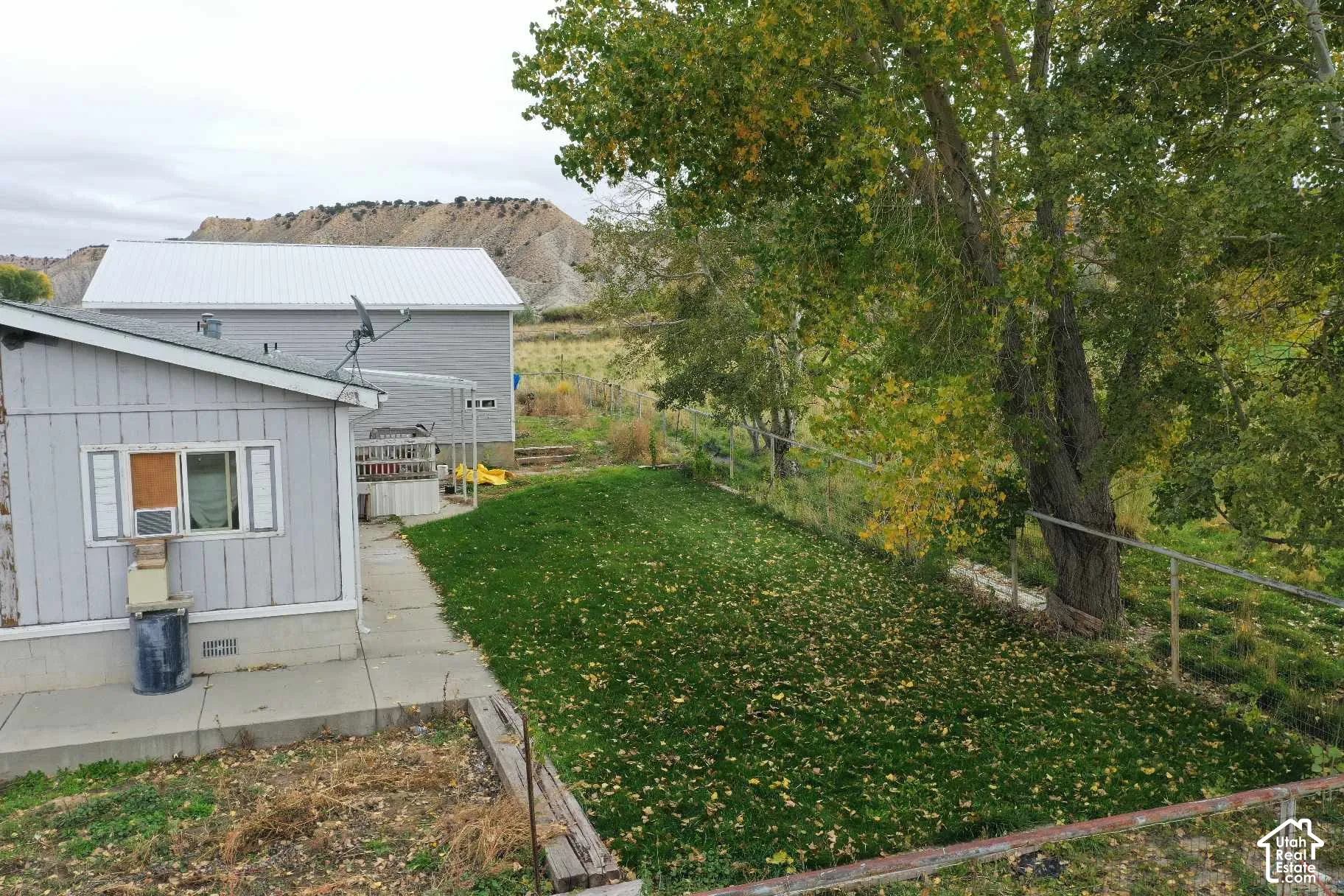 Exterior space featuring a concrete patio, grassy lawn, mature trees, and metal perimeter fencing against a backdrop of mountain terrain