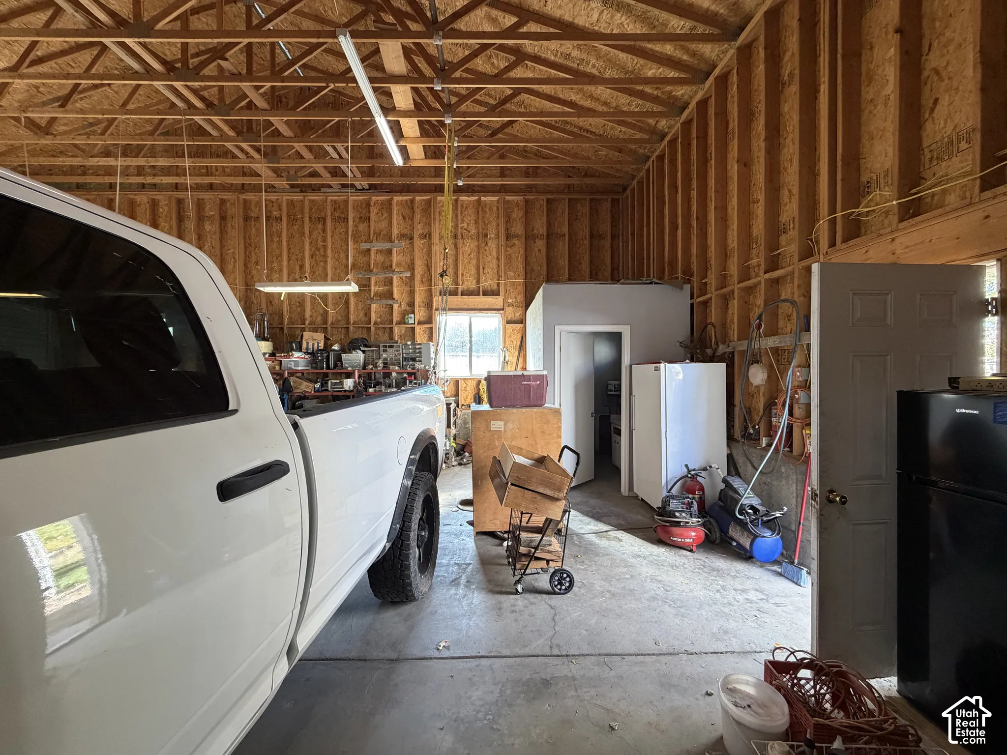 Spacious garage featuring exposed wood-stud framing, vaulted timber trusses, concrete flooring, linear fluorescent lighting, and an enclosed utility room