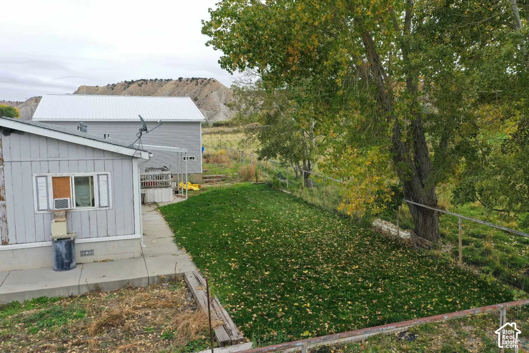 Exterior space featuring a concrete patio, grassy lawn, mature trees, chain-link fencing, and a mountain backdrop