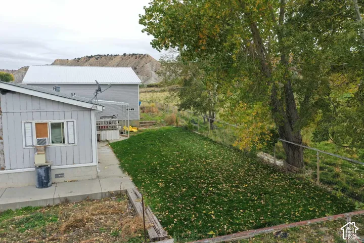 Exterior space featuring a concrete patio, grassy lawn, mature trees, and metal perimeter fencing against a backdrop of mountain terrain