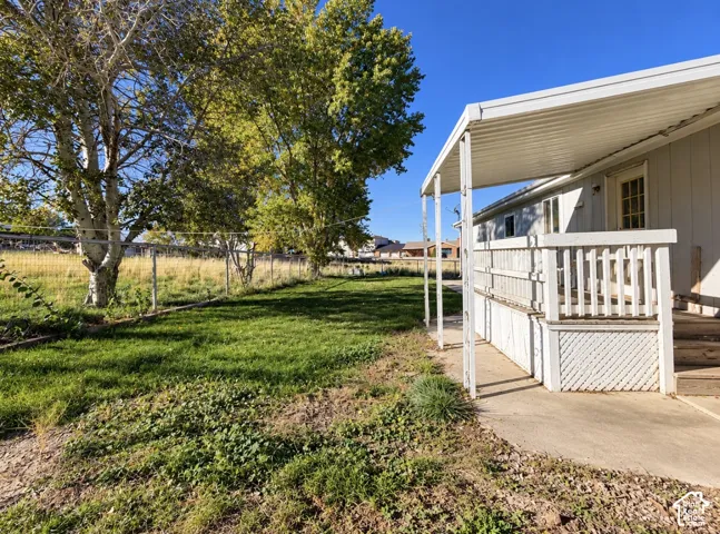 Exterior side yard featuring a covered porch with white railings, lattice skirting, concrete walkway, chain-link fencing, and mature trees