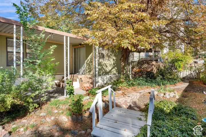 Covered patio featuring white structural support columns, stone-lined garden path, decorative wooden footbridge, mature trees, and landscape ground cover
