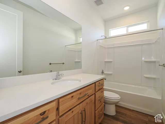 Bathroom featuring a wood-finish vanity with a light-colored solid surface countertop, an undermount sink, a full-width vanity mirror, a tub-shower combination with integrated shelving, and wood-finish flooring