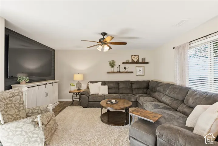 Living area featuring wood-finish flooring, a dark-toned shiplap accent wall, a central ceiling fan with integrated lighting, and horizontal window blinds