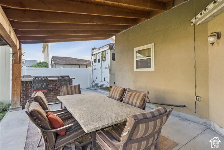 Covered patio featuring an exposed timber-beam ceiling, concrete flooring, exterior wall-mounted lighting, and a built-in brick outdoor kitchen island
