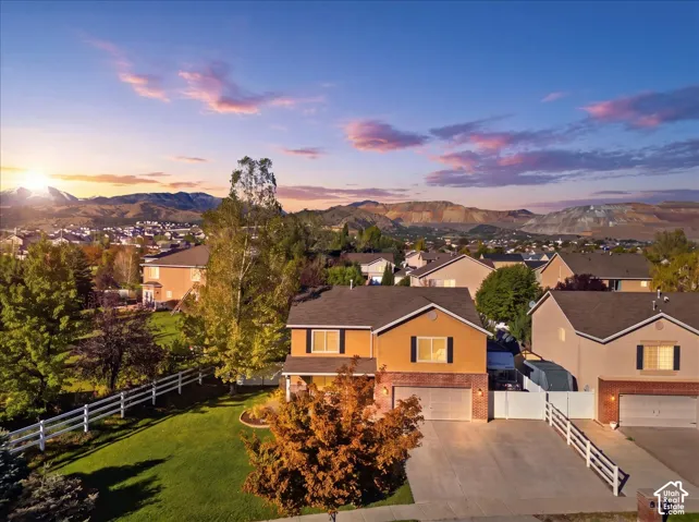 Two-story residence featuring a brick and stucco exterior, attached multi-car garage, concrete driveway, and expansive lawn area enclosed by white split-rail fencing, all set against a prominent mountain backdrop