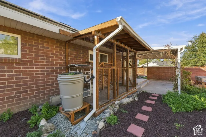 Exterior brick facade featuring a covered timber-frame structure with wire mesh screening, an integrated rainwater collection barrel, and a stone-lined concrete pathway